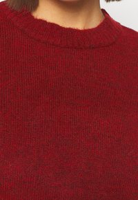 Punainen neulepaita, jossa on ribbikuvio ja klassinen pyöreä pääntie. Sujuva pinta, jossa on tasainen väri koko vaatteessa.