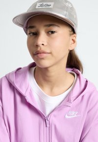 Felpa con zip rosa con logo Nike bianco, indossata sopra una maglietta bianca, abbinata a un cappellino argentato lucido con un'etichetta che dice "Lil Boo."