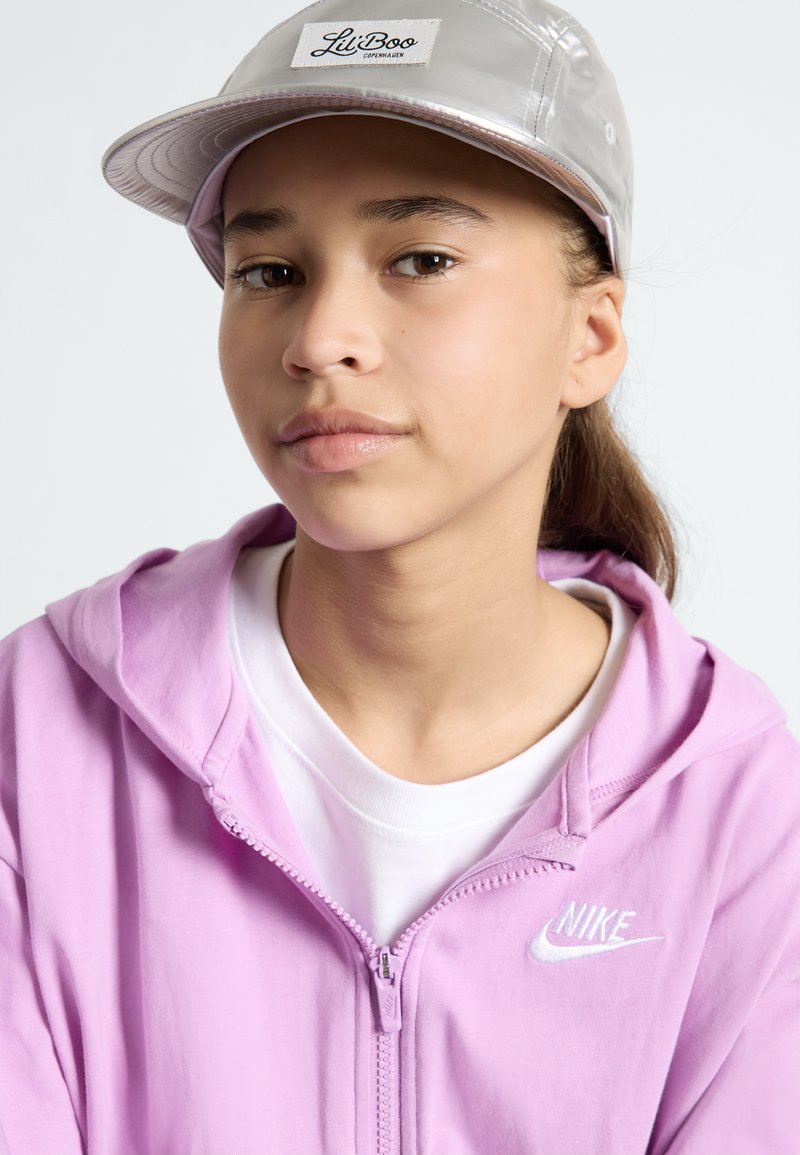 Felpa con zip rosa con logo Nike bianco, indossata sopra una maglietta bianca, abbinata a un cappellino argentato lucido con un'etichetta che dice "Lil Boo."
