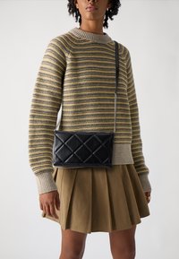 Svart quiltet crossover-veske med sølvkjede-stropp. Bæres med en stripete strikkegenser i gul, grå og beige, og en beige plisserte skjørt.