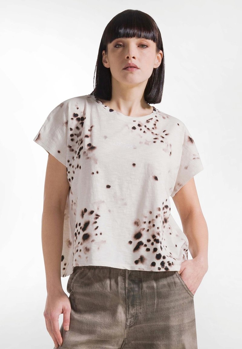 Femme aux cheveux noirs raides portant un t-shirt blanc ample avec des taches foncées et un pantalon marron, debout avec une main dans la poche.