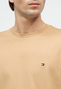 Κοντινό πλάνο του λαιμού και του στήθους ενός ατόμου που φοράει μπεζ T-shirt με στρογγυλή λαιμόκοψη και ένα μικρό κόκκινο, λευκό και μαύρο ορθογώνιο λογότυπο στην αριστερή πλευρά.