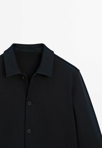 Cappotto nero con un collo a camicia, caratterizzato da una superficie liscia e chiusura con bottoni. Il design presenta dettagli minimi e spalle strutturate.