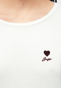 T-shirt cinza claro com decote redondo. Apresenta um gráfico de coração em bordô e texto manuscrito "Boys" no lado esquerdo do peito. Textura macia.