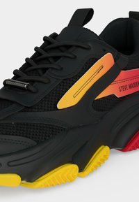 Μαύρο αθλητικό παπούτσι με κίτρινες και κόκκινες λεπτομέρειες στη σόλα, μαύρα κορδόνια, λεπτομέρειες από δίχτυ και την επωνυμία "Steve Madden" στο πλάι.