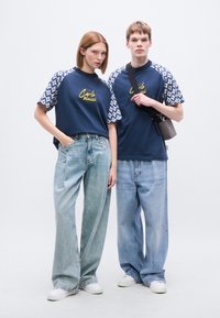 Μπλε μπλουζάκια ναυτικού με μανίκια με σχέδια, με κίτρινη κεντητή γραφή· άνετα ανοιχτό μπλε τζιν· μοντέλα που φορούν λευκά αθλητικά παπούτσια.