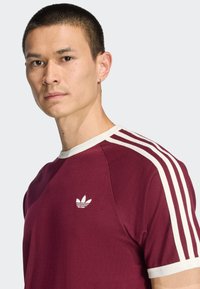 Μπλουζάκι σε βουργουνδί απόχρωση με λευκές λεπτομέρειες και τρεις κάθετες ρίγες στους ώμους. Διαθέτει ένα μικρό λευκό λογότυπο στο στήθος. Μαλακή υφή.