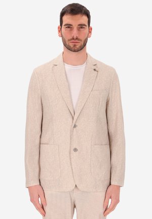 Uomo con capelli scuri e corti e barba, che indossa un blazer beige a due bottoni e pantaloni coordinati sopra una maglietta bianca a girocollo, in piedi davanti a uno sfondo bianco.