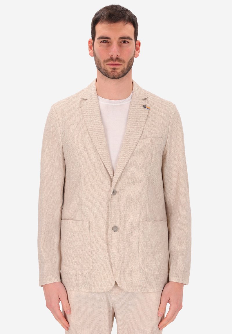 Uomo con capelli scuri e corti e barba, che indossa un blazer beige a due bottoni e pantaloni coordinati sopra una maglietta bianca a girocollo, in piedi davanti a uno sfondo bianco.