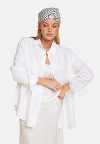 Hvit lin skjorte med lange ermer, lagd over en lysfarget tanktop, parret med et mønstret hodeplagg med striper.