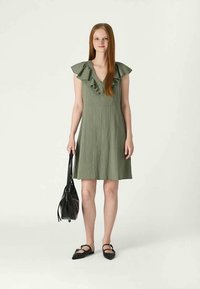 Πράσινο, μίνι φόρεμα μέχρι το γόνατο με ριχτές μανίκες και υφασμάτινη υφή. Συνδυασμένο με μαύρες φλατ παπούτσια και τσάντα ώμου. Απλό φόντο.