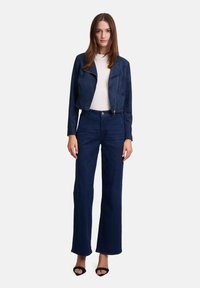 Marineblaues Wildleder-Cropped-Jackett mit einem Reißverschluss, kombiniert mit hoch taillierten, dunkelblauen Jeans im Schlag und schwarzen offenen High Heels. Darunter ein weißes Shirt.