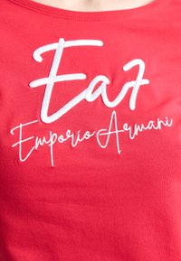 Sarkans kokvilnas T-krekls ar baltu izšuvumu "EA7 Emporio Armani" logotipa tekstu, kas veidots apvienojot treknrakstu un itāļu slēdzienu. Gluda tekstūra.