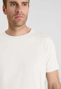 Uomo con leggera barba incolta che indossa una semplice t-shirt bianca a maniche corte con scollo rotondo, che guarda leggermente a sinistra su uno sfondo grigio neutro.