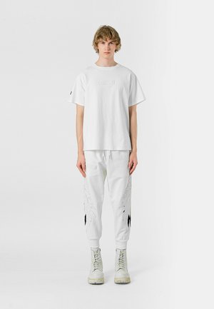 Camiseta de algodón blanca con un parche cuadrado, combinada con joggers blancos que cuentan con bordados en forma de alas y detalles en negro, completada con botas blancas.