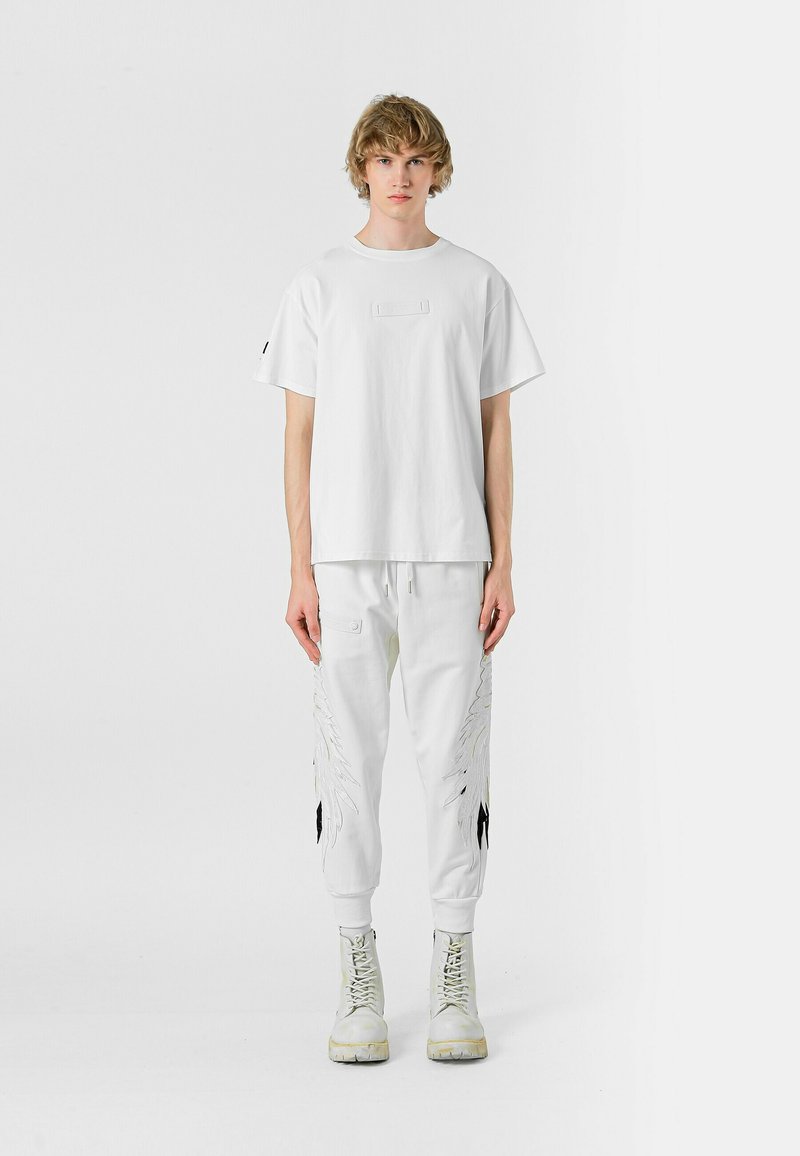 Camiseta de algodón blanca con un parche cuadrado, combinada con joggers blancos que cuentan con bordados en forma de alas y detalles en negro, completada con botas blancas.