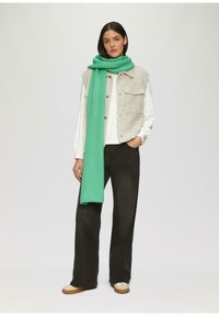 Lichtgrijs structuurvest over een witte blouse, gecombineerd met zwarte brede broek en een felgroene sjaal, afgewerkt met crèmekleurige en bruine schoenen.