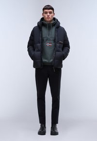 Giacca a vento nera con cappuccio, felpa verde con design della bandiera, pantaloni neri e stivali neri. Tessuto elegante con una superficie liscia.