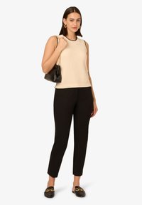 Beige gestricktes ärmelloses Oberteil mit schwarzem Rand, kombiniert mit engen schwarzen Hosen und schwarzen Loafers mit goldenen Akzenten, gehalten mit einer schwarzen Handtasche.