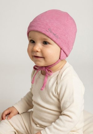 Kleinkind trägt eine rosa gestrickte Mütze mit Bändern, bekleidet mit einem cremefarbenen Langarmshirt und einer Hose, sitzt und lächelt vor einem schlichten Hintergrund.