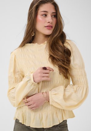 Jeune femme aux longs cheveux ondulés, portant un chemisier jaune pâle à manches bouffantes et des bijoux en or, regardant sur le côté sur un fond uni.