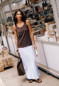 Femme portant un haut marron sans manches et une jupe blanche, tenant un livre et un sac fourre-tout, debout dans une allée avec des livres empilés sur des étagères.