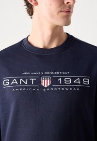 Tmavě modrá mikina s kulatým výstřihem, na přední straně s grafickým potiskem, na kterém je text a štítové logo v červené a bílé barvě.