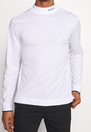 Bílé bavlněné triko s dlouhým rukávem a stojáčkem, obtaženého střihu. Na límci je černé logo a na manžetách námořnicky modré detaily.