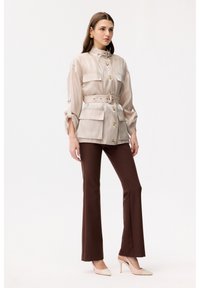Woman wearing beige belted jacket with large pockets, dark brown flared pants, beige pointed high heels, and gold earrings, standing against white background.