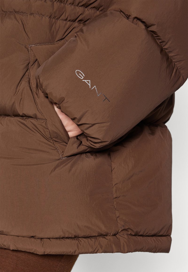 Brązowa kurtka puchowa o błyszczącym wykończeniu, z kieszonką boczną i logo "GANT" w srebrnym hafcie na rękawie.