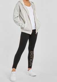 Lichtgrijze zip-up hoodie met witte strepen, witte tanktop, zwarte leggings met "Bench" logo en witte sportschoenen.