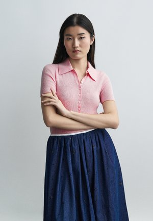 Νεαρή γυναίκα με μακριά μαύρα μαλλιά, ντυμένη με ροζ μπλούζα με κουμπιά και κοντά μανίκια και μπλε φούστα με μικρά κεντητά σχέδια.