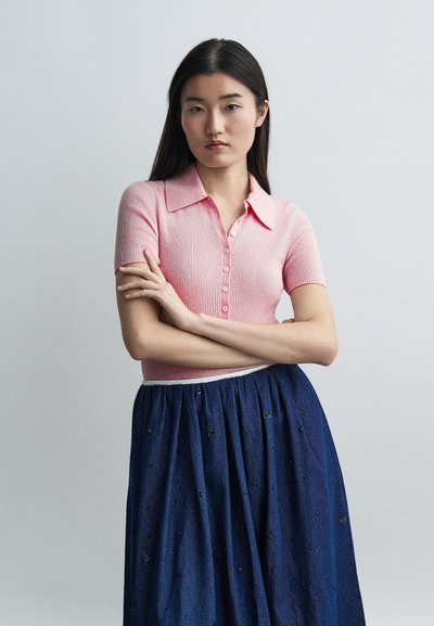 Jeune femme aux longs cheveux noirs portant un haut rose à manches courtes boutonné et une jupe bleue ornée de petits motifs brodés.