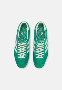 Gröna mockaskor med vit tre-stripes-design, vita snören och vadderad tunga. Interiören har ett Adidas-logomönster i grönt.