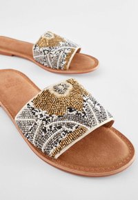 Perlenbesetzte Schippersandalen mit einem breiten Riemen, der goldene, silberne und schwarze Perlen in Blumenmustern zeigt, sowie einer Fußbett aus braunem Wildleder.