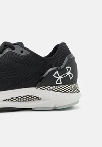 Zwarte sportieve schoen met een bovenkant van mesh, gestructureerde stof, witte zool met een meshpatroon en een glanzende zwarte accent met een logo.