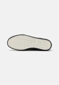 Zapatillas negras con una suela de goma texturizada, que presentan un patrón de zigzag blanco y un tacón moderado. Branding mínimo en el lateral.