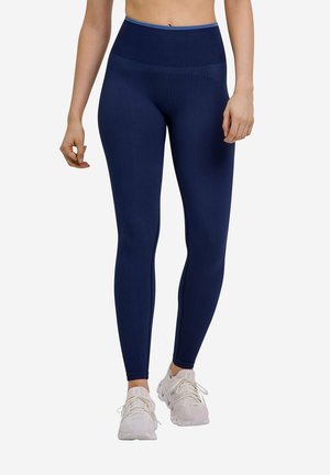 Frau trägt marineblaue Leggings mit hoher Taille und weiße Laufschuhe und steht vor einem schlichten Hintergrund.