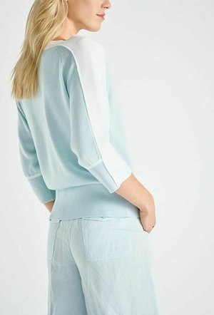 Femme aux longs cheveux blonds portant un pull bleu clair avec des panneaux blancs aux épaules et un pantalon bleu clair assorti, debout les mains dans les poches.