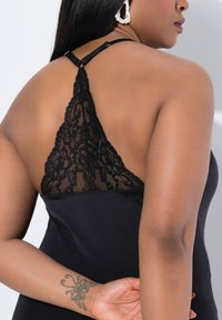 Abito nero con design in pizzo sulla schiena, dotato di spalline regolabili e una texture liscia. Include un tatuaggio visibile sul braccio sinistro.