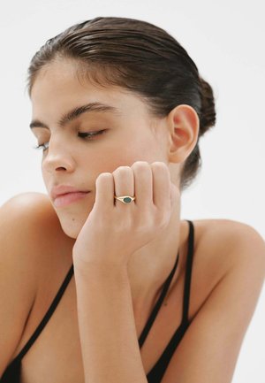 Anillo de color dorado con una piedra verde ovalada, llevado en el dedo índice. El fondo es blanco liso, destacando el diseño del anillo.