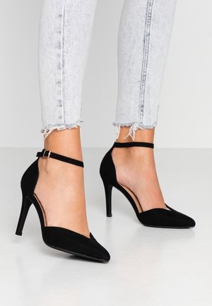 Sorte ruskinds højhælede sko med spidse tåer og ankelrem. Har et udskåret design og en slank stiletto-hæl.