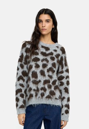 Kobieta w szarym, puszystym swetrze z dużymi ciemnobrązowymi cętkami w panterkę oraz niebieskich dżinsach, stojąca na tle jednolitego, jasnego tła.