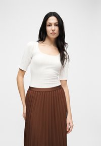 Γυναίκα με μακριά σκούρα μαλλιά, φορούν λευκή μπλούζα με στρογγυλή λαιμόκοψη και καφέ πλισέ φούστα με ψηλή μέση, στέκεται μπροστά σε απλό ανοιχτό φόντο.
