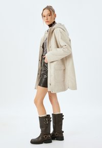 Abrigo de mezcla de lana beige con capucha, botones a presión y bolsillos delanteros, combinado con una camisa de cuadro, falda de cuero sintético negra y botas marrones con hebilla.