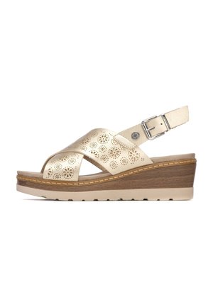Sandalia de cuña de cuero dorado con tiras cruzadas, diseño floral perforado, hebilla ajustable y suela beige texturizada.