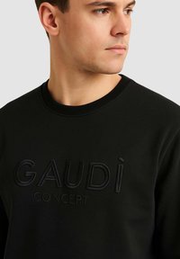 Felpa nera con scollo a coste, caratterizzata da testo in rilievo "GAUDÌ CONCEPT" in tono nero abbinato per un look e una texture coerenti.