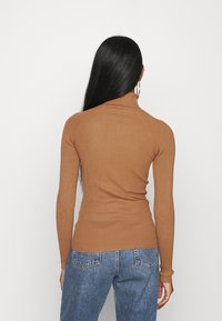 Kobieta z długimi czarnymi włosami, ubrana w dopasowany brązowy prążkowany sweter z golfem i niebieskie dżinsy, stoi tyłem na jednolitym tle.