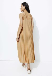 Robe longue plissée de couleur champagne avec un dos en V et des bretelles fines, associée à des sandales à talons métalliques. Texture lisse et silhouette fluide.