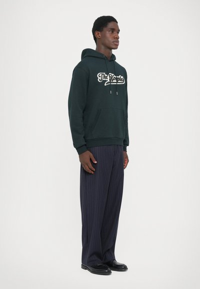Green hoodie with white text logo, kangaroo pocket, drawstring hood; paired with navy pinstriped trousers and black shoes.